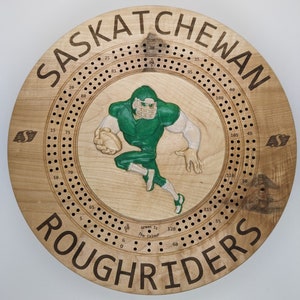 Puede incluir: Un tablero de juego de madera con un jugador de fútbol verde y blanco en el centro. El tablero es circular y tiene una serie de agujeros alrededor del borde. El texto "SASKATCHEWAN ROUGHRIDERS" está impreso alrededor del borde del tablero.