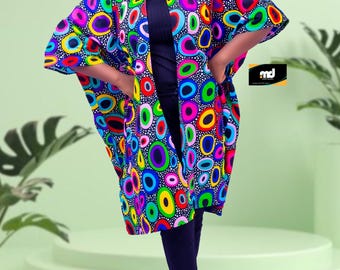 May include: A colourful, patterned kimono-style jacket with an abstract design of overlapping circles in various colours. The jacket is worn over a black turtleneck and leggings. White trainers complete the outfit.