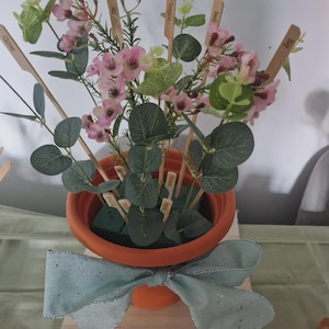 May include: A terracotta flower pot filled with pink flowers, green leaves, and wooden herb markers. A light blue bow with silver glitter accents adorns the pot. The wooden markers are labelled with herb names.