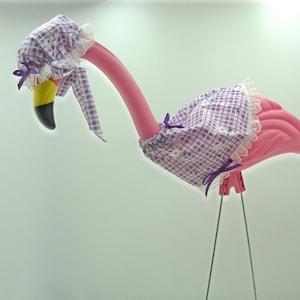 May include: A whimsical pink flamingo statue with a black and yellow beak, adorned with a purple and white checkered bonnet and dress, trimmed with white lace and purple bows. The flamingo stands on thin metal legs.