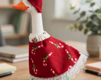 May include: A white goose figurine wearing a red felt hat with green leaves and a red dress featuring a candy cane and holly pattern. The dress has a white ruffled collar and a white lace trim along the bottom hem. The goose has an orange beak.