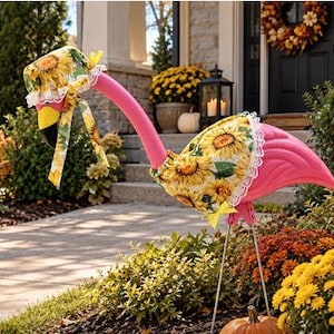 May include: A pink plastic flamingo lawn ornament wearing a sunflower-patterned fabric bonnet with white lace trim and a matching sunflower-patterned fabric cape with white lace trim, standing on two thin metal legs in a garden setting.