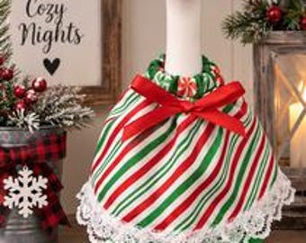 May include: A white goose figurine wearing a red and green striped fabric cape with a red ribbon bow at the neck, a white lace trim along the bottom edge, and a small red and white hat with green accents on its head, standing in a festive display.