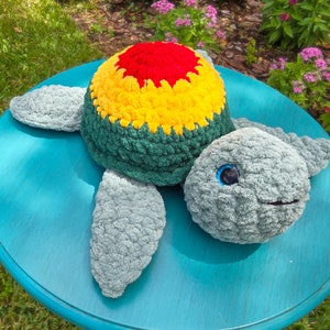Puede incluir: Un peluche de tortuga hecho a mano. El caparazón es rojo, amarillo y verde. La cabeza y las aletas son grises. Tiene una cara sonriente y ojos azules. Se encuentra sobre una superficie azul turquesa.