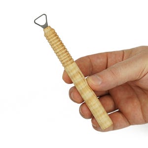 May include: A pottery trimming tool featuring a light-colored wooden handle with a ribbed grip section and a small, triangular metal loop at the tip. The handle has a natural wood grain pattern and a smooth, cylindrical shape for manual use.