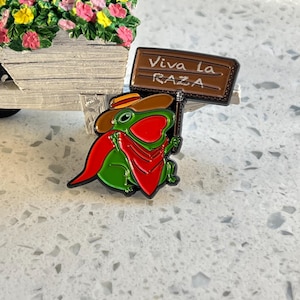 May include: A green frog pin wearing a cowboy hat and red scarf, holding a sign that reads "Viva La Raza." The pin is set against a white surface, with a miniature wagon filled with flowers in the background.