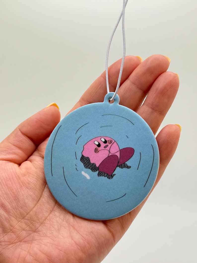 K&ouml;nnte beinhalten: Ein runder blauer Auto-Lufterfrischer mit einer pinkfarbenen Comicfigur in der Mitte. Die Figur ist von einem blauen Kreis mit wei&szlig;en Wellen umgeben. Der Lufterfrischer h&auml;ngt an einer wei&szlig;en Schnur.