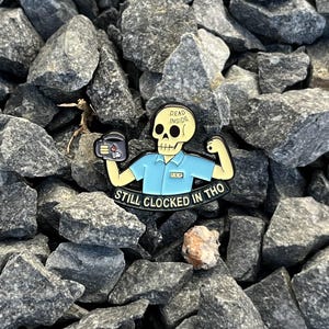 May include: A black and yellow enamel pin featuring a skeleton in a blue shirt holding a coffee mug. The pin reads "DEAD INSIDE" and "STILL CLOCKED IN THO". The pin is set against a background of gray rocks.