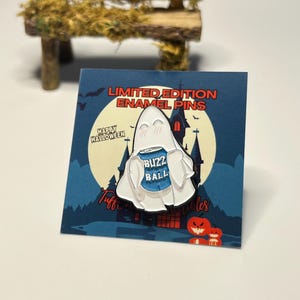 May include: A white ghost enamel pin with a blue "Buzz Ball" can in its hands. The pin is on a blue and white card with the text "Limited Edition Enamel Pins" and "Happy Halloween".