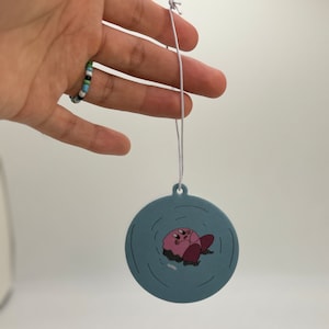 K&ouml;nnte beinhalten: Ein runder blauer Auto-Lufterfrischer mit einer pinkfarbenen Comicfigur in der Mitte. Die Figur schwebt im Wasser mit Wellen darum herum.
