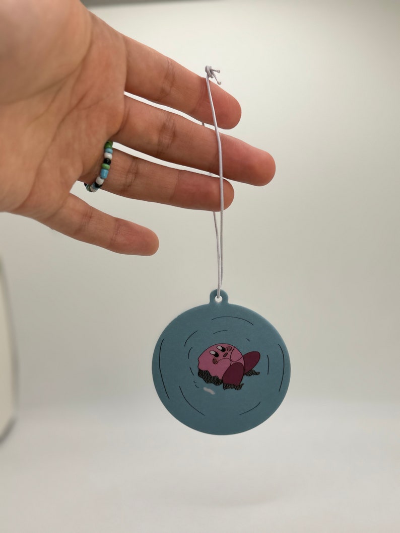 K&ouml;nnte beinhalten: Ein runder blauer Auto-Lufterfrischer mit einer pinkfarbenen Comicfigur in der Mitte. Die Figur schwebt im Wasser mit Wellen darum herum.