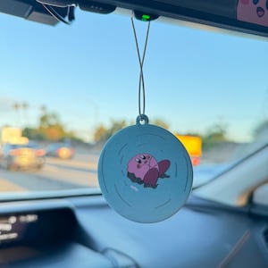 K&ouml;nnte beinhalten: Ein rosa-blauer Auto-Lufterfrischer mit einer Comicfigur in der Mitte. Die Figur ist rosa und hat einen runden K&ouml;rper mit einem kleinen Mund und Augen. Die Figur schwebt in einem blauen Kreis mit wei&szlig;en Wellen darum herum.