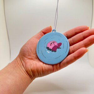 K&ouml;nnte beinhalten: Ein runder blauer Auto-Lufterfrischer mit einer pinkfarbenen Comicfigur in der Mitte. Die Figur schwebt in einem blauen Kreis mit wei&szlig;en Wellen.