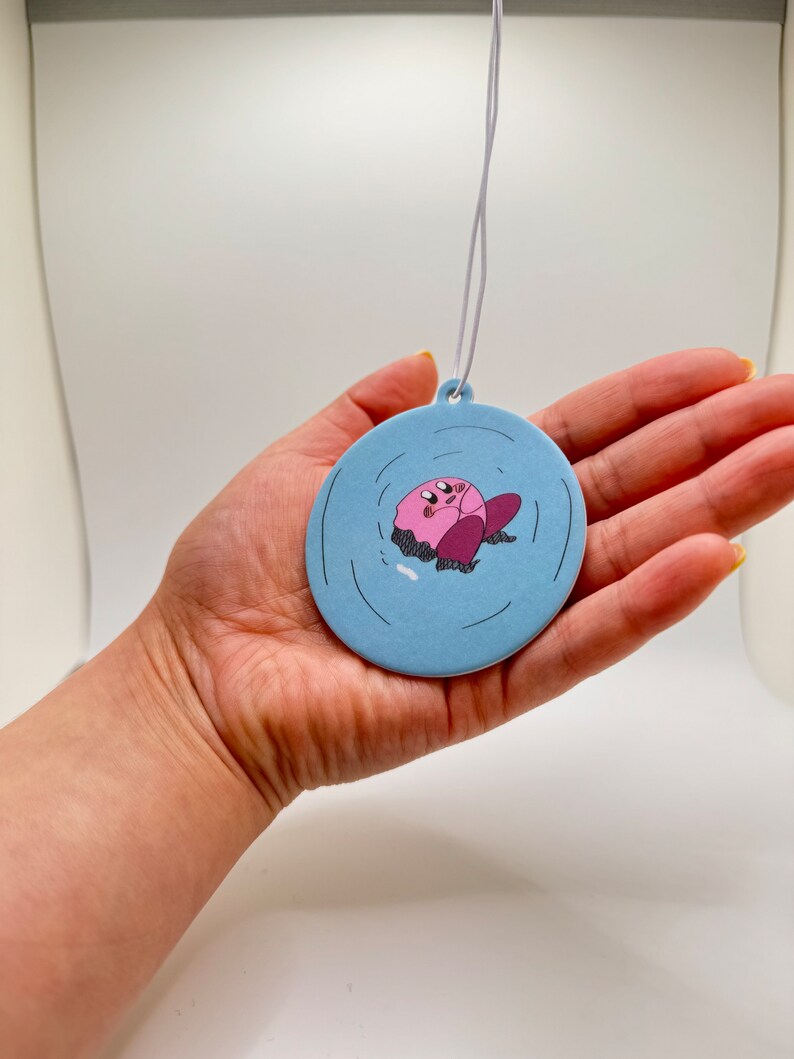 K&ouml;nnte beinhalten: Ein runder blauer Auto-Lufterfrischer mit einer pinkfarbenen Comicfigur in der Mitte. Die Figur schwebt in einem blauen Kreis mit wei&szlig;en Wellen.