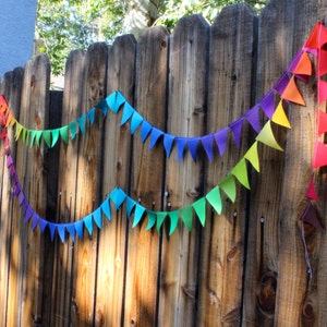 Peut inclure: Une bannière en feutre arc-en-ciel avec des drapeaux triangulaires suspendus à une clôture en bois. La bannière est faite de différentes nuances de rouge, orange, jaune, vert, bleu, indigo et violet.