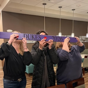 May include: Three people hold a purple wooden sign with the text "DUKES 2026" in black lettering. The sign has red circular accents on each end.