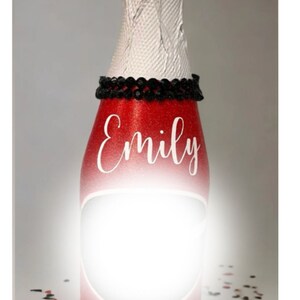 May include: A red and white mini champagne bottle with a black beaded trim and the name "Emily" written in white. The bottle is sitting on a white surface with red and black confetti.
