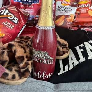 May include: A red and gold bottle with a glitter finish and a white design that says "Lafayette" and "Madeline". The bottle is surrounded by a black sweatshirt with white text, leopard print slippers, and bags of chips.