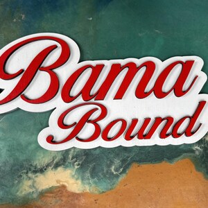 May include: White wooden sign with the words "Bama Bound" in red cursive lettering.