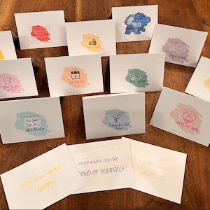 May include: A set of 12 cards with colorful backgrounds and encouraging messages. Each card has the text "Open when you are..." followed by a positive message, such as "Happy", "Proud of Yourself", or "Out of Funds".