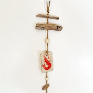 May include: A vertical wall hanging featuring two pieces of driftwood, two light-colored wooden beads, a rectangular ceramic tile with a red shrimp illustration, and a small black circular disc, all strung together on a tan cord.