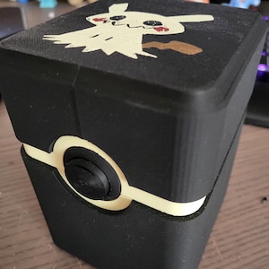 May include: A black, rectangular box with a cream-colored accent and a circular button. The top of the box features a cream-colored cartoon ghost design with red and brown accents. The box is likely for storage.