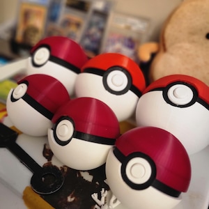 May include: A collection of red, white, and black Poké Ball replicas. The spherical objects have a white base, a red top, and a black band around the middle. A white circle is centered on the front of each ball.