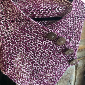 May include: A purple and white knitted scarf with wooden buttons. The scarf is draped over a hanger and has a loose, open weave.