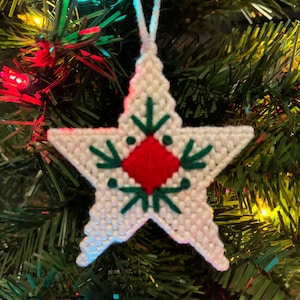May include: White star-shaped Christmas ornament with a red and green embroidered design. The ornament is hanging on a green Christmas tree.