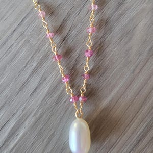 May include: A delicate gold necklace featuring pink gemstone beads and a large, pear-shaped white pearl pendant. The necklace has a fine chain and is displayed against a neutral gray background.