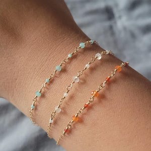 May include: Three delicate gold chain bracelets adorned with small, colorful beads. One bracelet features pale blue beads, another has white beads, and the third is accented with orange beads. The bracelets are worn on a wrist.