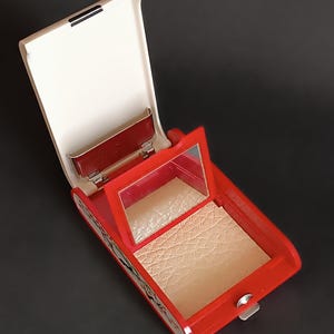 K&ouml;nnte beinhalten: Ge&ouml;ffnete rote und cremefarbene Kompaktpuder-Dose mit Spiegel und Puderfach. Die rechteckige Dose hat abgerundete Kanten und einen silbernen Verschluss. Das Puder ist hellbeige. Der Spiegel ist rechteckig.