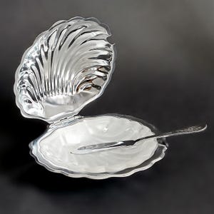 May include: A silver-toned metal clam shell-shaped dish with a hinged lid that opens upward. The interior of the dish features a smooth, white, scalloped surface. A matching silver-toned metal butter knife rests horizontally inside the open shell.