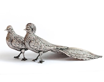 May include: Two silver pheasant figurines with detailed feather patterns. The pheasants are standing on a white surface.