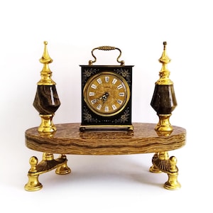 Könnte beinhalten: Eine antike Messinguhr mit schwarzem Zifferblatt und goldenen Zeigern, die auf einem Marmorsockel mit zwei dekorativen Messingzieraten steht.