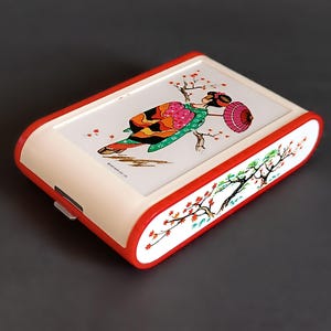 K&ouml;nnte beinhalten: Eine rechteckige, cremefarbene Box mit rotem Rand. Die Oberseite zeigt eine farbenfrohe Illustration einer Geisha mit Sonnenschirm und Kirschbl&uuml;ten. Die Seiten sind mit einem Kirschbl&uuml;tenzweig verziert. Ein kleiner Metallverschluss ist sichtbar.