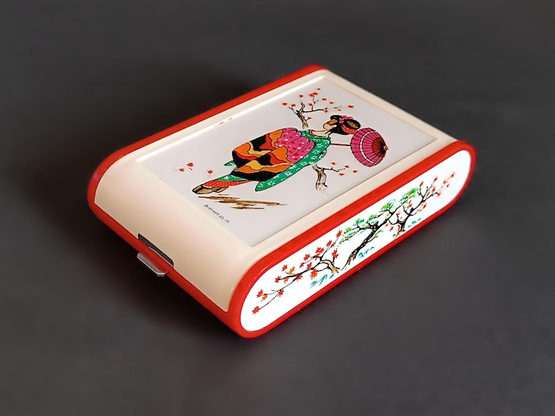 K&ouml;nnte beinhalten: Eine rechteckige, cremefarbene Box mit rotem Rand. Die Oberseite zeigt eine farbenfrohe Illustration einer Geisha mit Sonnenschirm und Kirschbl&uuml;ten. Die Seiten sind mit einem Kirschbl&uuml;tenzweig verziert. Ein kleiner Metallverschluss ist sichtbar.