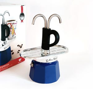 Peut inclure: Une cafetière italienne bleue et argentée avec une poignée noire. La cafetière a un dessus argenté avec deux becs courbés et une base bleue octogonale. Une boîte avec une tasse à café est visible en arrière-plan.