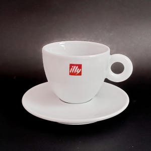 May include: White ceramic coffee cup and saucer set with the word "illy" in red lettering on the cup.