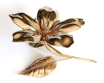 May include: A gold-colored metal flower sculpture with detailed petals, a central stamen, a stem, and a leaf. The sculpture is set against a white background, highlighting its intricate design and metallic sheen. The flower is a magnolia.