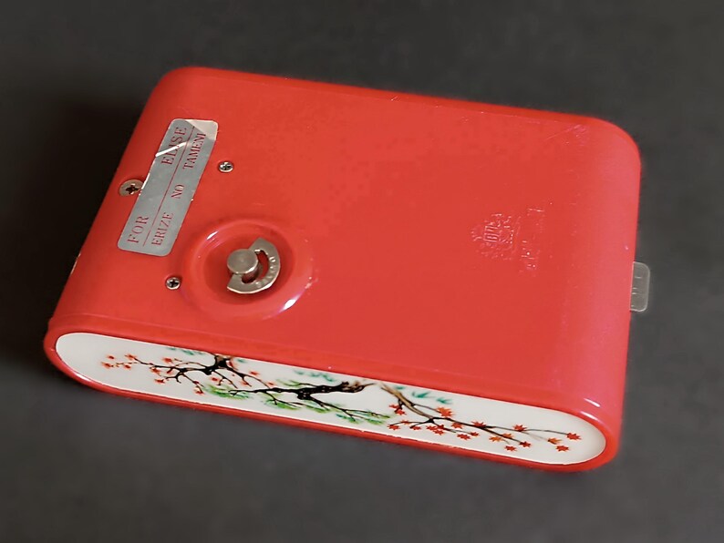 K&ouml;nnte beinhalten: Eine rote Vintage-Kamera mit floralem Design am unteren Rand. Die Kamera hat einen silbernen Aufzugsknopf und ein Etikett mit der Aufschrift "FOR ERIZE NO TAMEW ELSE".