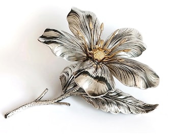 May include: A silver-toned flower brooch with gold-colored accents. The brooch features detailed petals, a central stamen, and a stem with a leaf. The design is intricate and elegant, suitable for formal wear or as a decorative accessory.