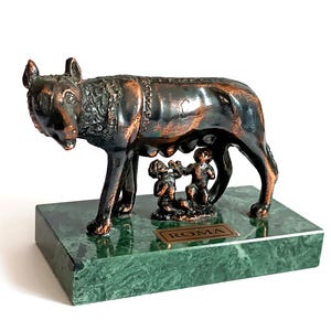 May include: A bronze sculpture of the Capitoline Wolf nursing Romulus and Remus, set on a green marble base. The wolf is dark brown with detailed fur, and the twins are small figures. A plaque on the base reads "ROMA".
