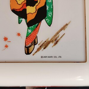 K&ouml;nnte beinhalten: Ein wei&szlig;es rechteckiges Tablett mit rotem Rand zeigt eine farbenfrohe Illustration einer Geisha in einem Kimono. Der Kimono der Geisha ist orange, gelb, gr&uuml;n und schwarz. Das Tablett tr&auml;gt den Text "LADY-MATE CO., LTD."
