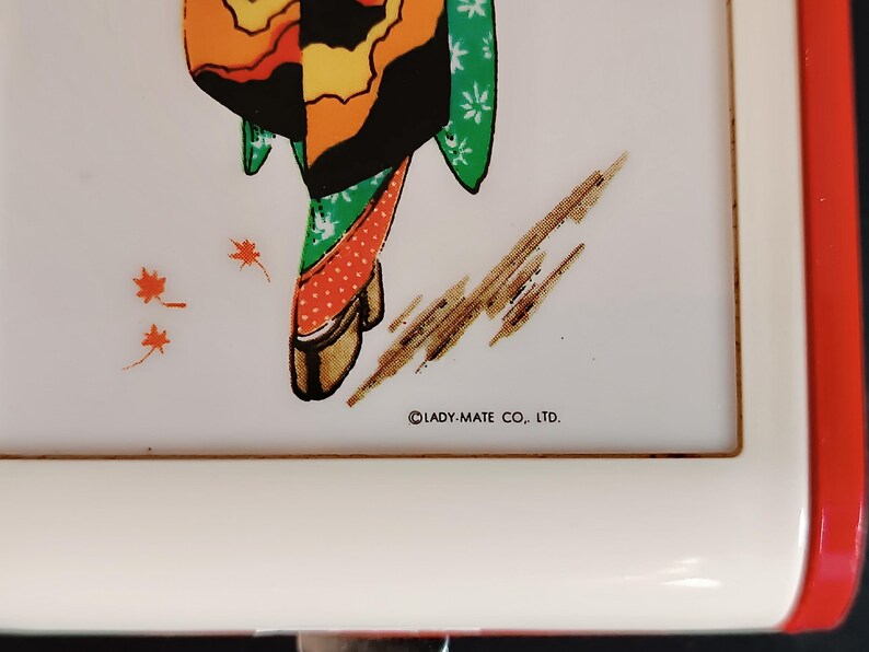 K&ouml;nnte beinhalten: Ein wei&szlig;es rechteckiges Tablett mit rotem Rand zeigt eine farbenfrohe Illustration einer Geisha in einem Kimono. Der Kimono der Geisha ist orange, gelb, gr&uuml;n und schwarz. Das Tablett tr&auml;gt den Text "LADY-MATE CO., LTD."