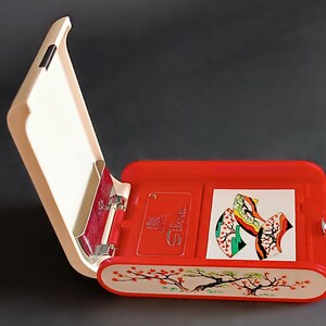 K&ouml;nnte beinhalten: Ge&ouml;ffnete rote und cremefarbene H&uuml;lle mit Klappdeckel. Die H&uuml;lle hat ein dekoratives Kirschbl&uuml;ten-Design am unteren Rand. Im Inneren befinden sich eine kleine Karte und eine farbenfrohe Illustration. Die H&uuml;lle ist ge&ouml;ffnet und gibt den Blick auf das Innere frei.