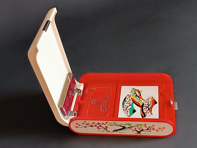 K&ouml;nnte beinhalten: Ge&ouml;ffnete rote und cremefarbene H&uuml;lle mit Klappdeckel. Die H&uuml;lle hat ein dekoratives Kirschbl&uuml;ten-Design am unteren Rand. Im Inneren befinden sich eine kleine Karte und eine farbenfrohe Illustration. Die H&uuml;lle ist ge&ouml;ffnet und gibt den Blick auf das Innere frei.