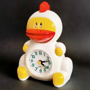 Op de afbeelding: Een witte en gele kwarts wekker in de vorm van een eend met een rode snavel en kam. De wijzerplaat heeft zwarte cijfers en groene accenten. De wijzers zijn zwart en geel.