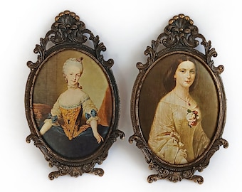 May include: Two antique oval portrait frames with ornate bronze-colored detailing. Each frame holds a vintage portrait of a woman. One portrait features a woman in a blue and gold dress, the other in a cream-colored dress.