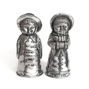 Peut inclure: Deux figurines argentées d'enfants en vêtements d'hiver. L'une porte un chapeau et un manteau, l'autre un chapeau, une écharpe et un manteau. Les deux ont des visages détaillés et des manteaux boutonnés, sur fond blanc.